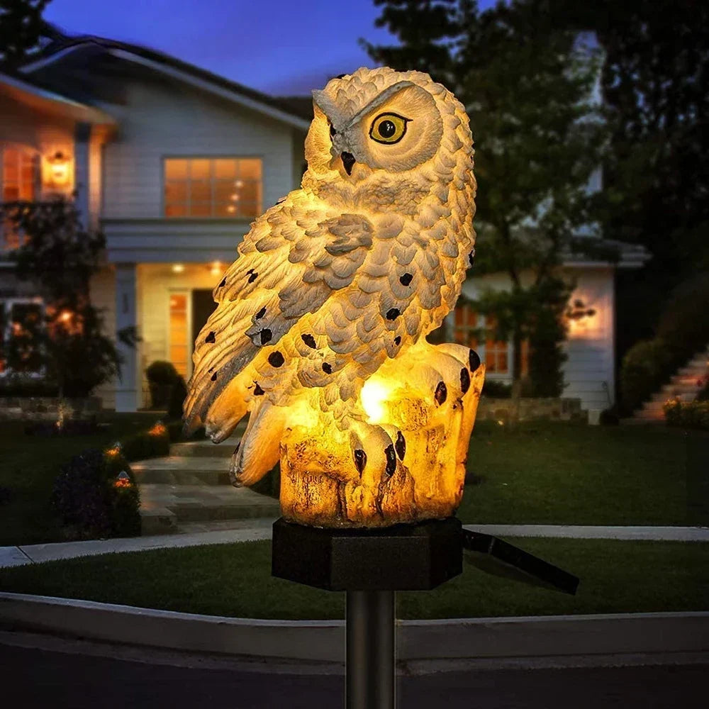 Weiße Eulenfigur mit warmem Licht am Baumstumpf-Sockel vor einem Haus.