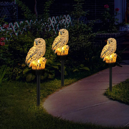 Drei Eulen-Gartenlichter auf Stäben am geschwungenen Gartenweg zwischen Büschen.