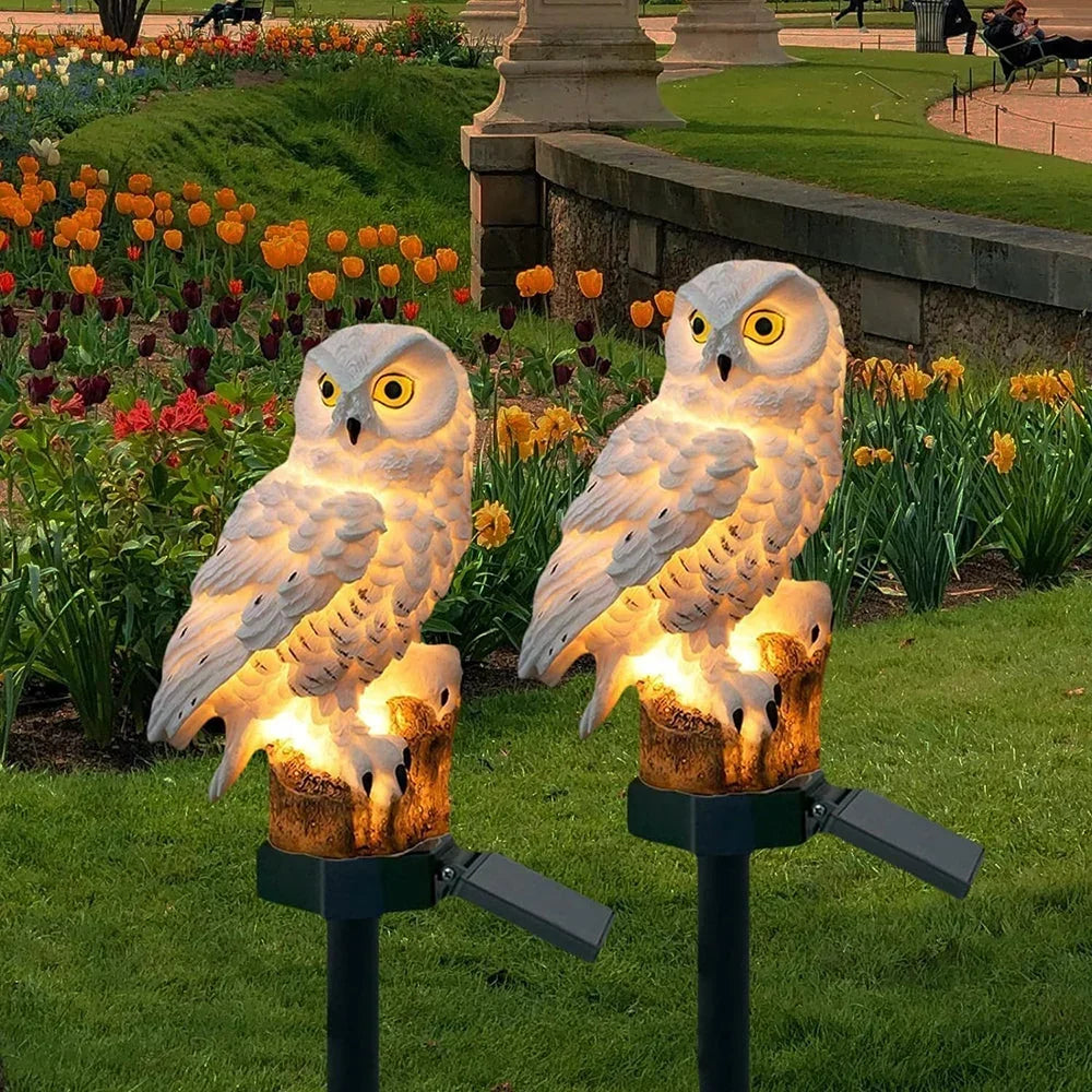 Zwei weiße Eulenfiguren mit Solarpanel auf Erdspießen im Rasen vor einem Tulpenbeet.