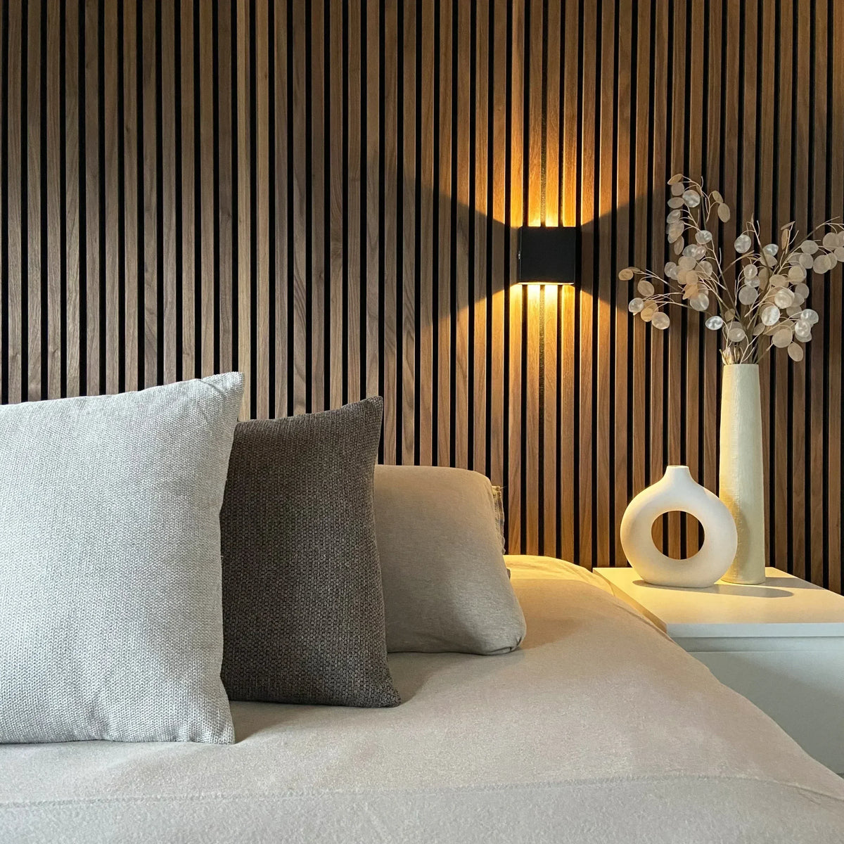 Schwarze Wandlampe vor einer Holzlamellenwand über einem Bett, warmes Licht, Kissen und weiße Vasen auf dem Nachttisch. 5