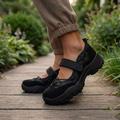 Schwarze Slipper mit Klettriemen auf einem Holzweg im Garten, getragen zu khakifarbener Jogginghose.