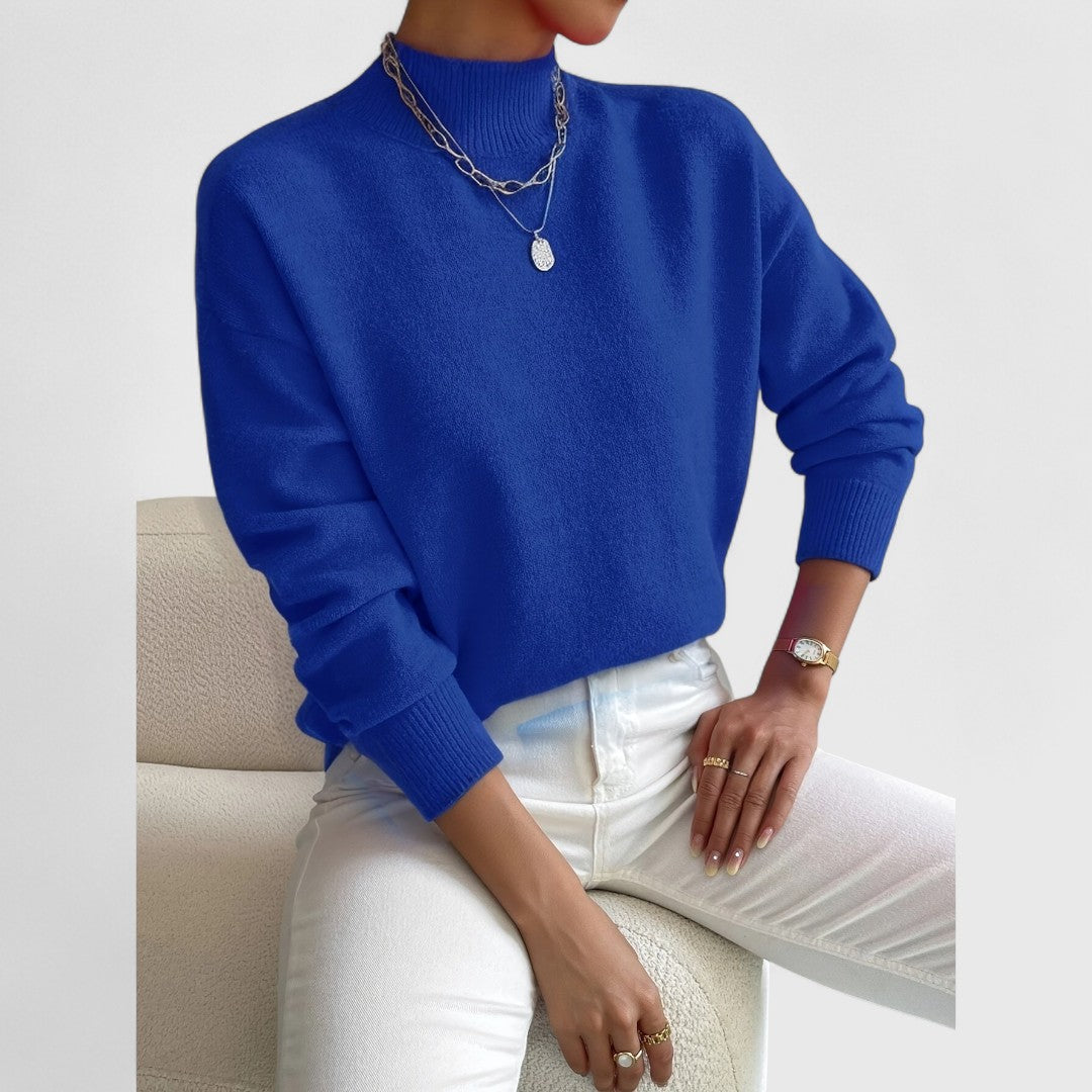 Damen Pullover in Königsblau mit Polokragen und weißer Jeans, sitzend auf einem Bouclé-Sessel.
