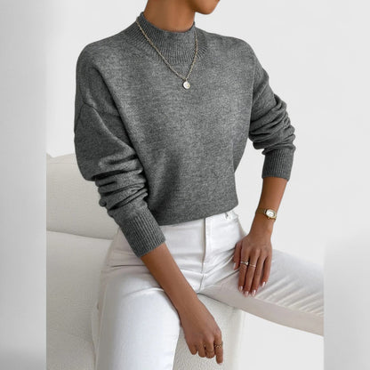 Damen Pullover in Grau meliert mit Polokragen und weißer Jeans, sitzend auf einem Sessel.