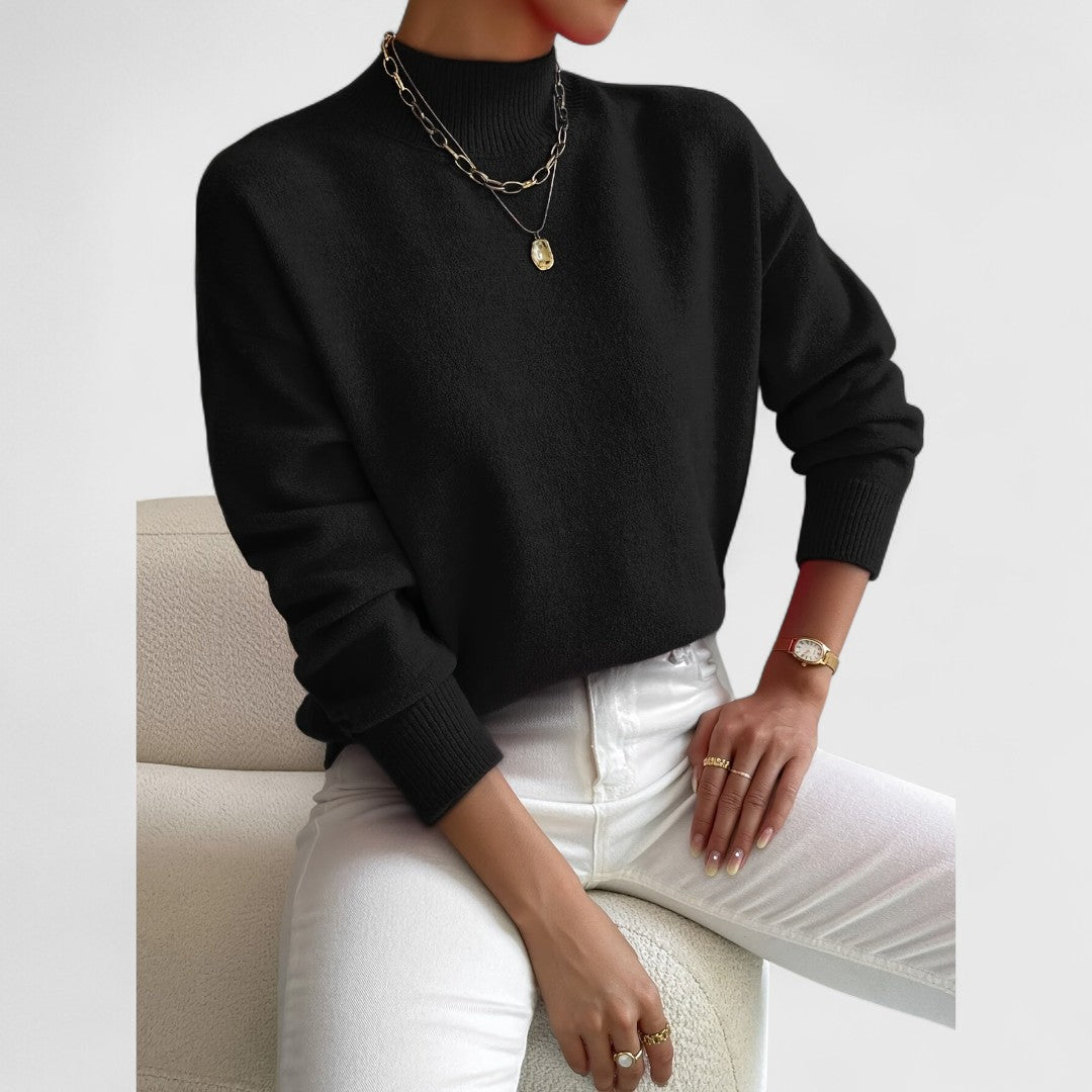 Damen Pullover in Schwarz mit Polokragen und weißer Jeans, sitzend auf einem Sessel.