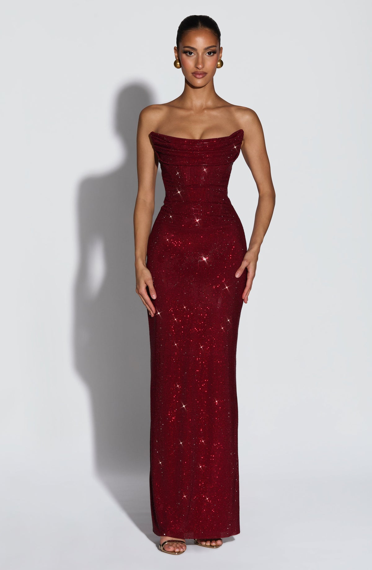 Model trägt das trägerlose Kleid in Dunkelrot, Vorderansicht mit Strasssteinen, drapiertem Oberteil und goldenen Ohrringen. 1