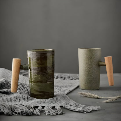 Zwei Becher mit Holzgriff, grün-braun glasiert und sandfarben, auf grauem Fransentuch neben einer Ähre 1
