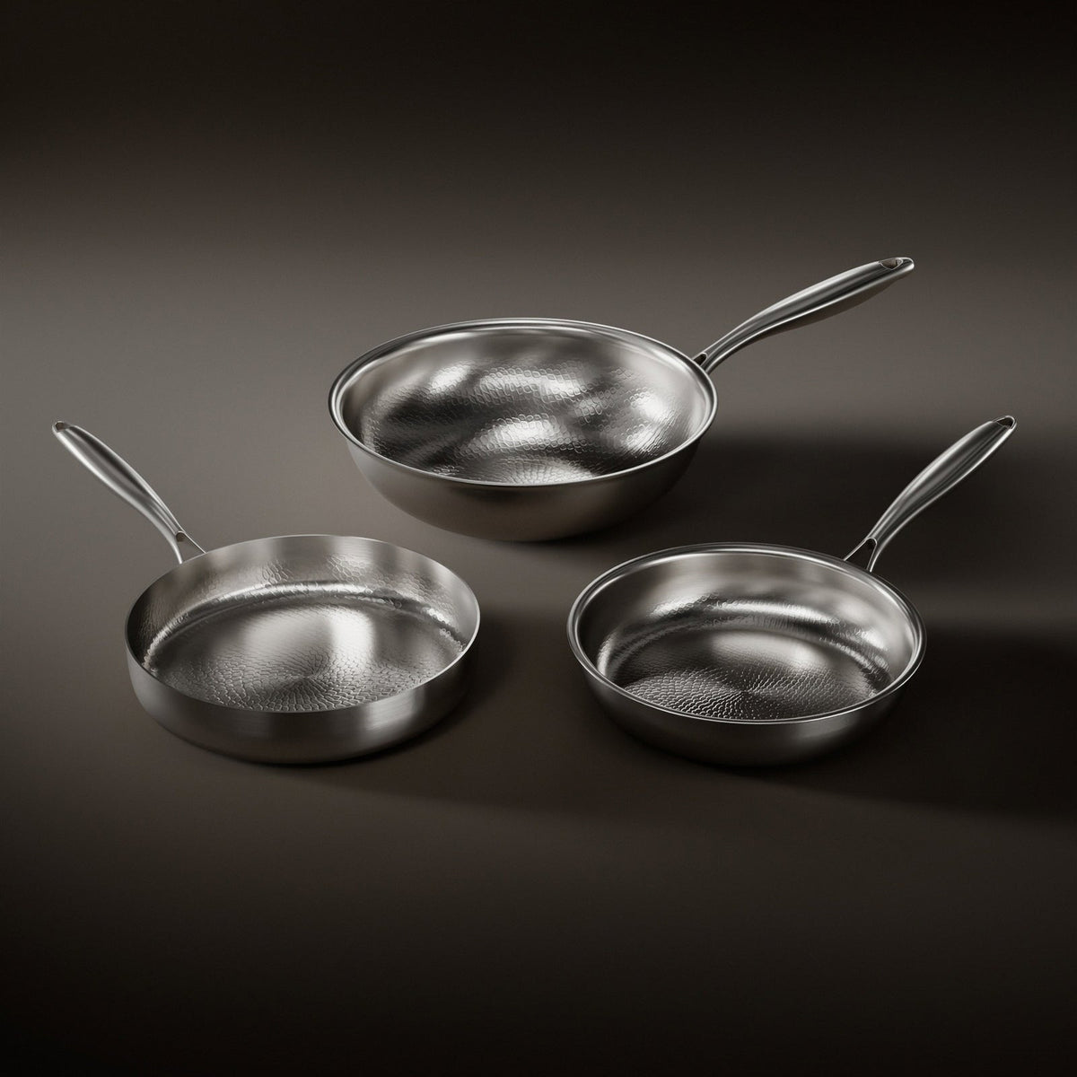 Drei Pfannen mit gehämmerter Innenfläche und Edelstahlgriffen, eine tiefe Pfanne, ein Wok und eine flachere Pfanne auf dunklem Hintergrund.