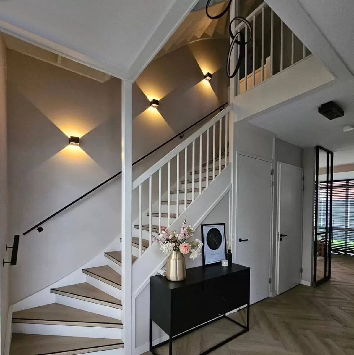 Drei schwarze Würfel-Wandlampen entlang einer Treppe im Flur, dazu schwarzes Sideboard mit Blumenvase und Bilderrahmen. 7