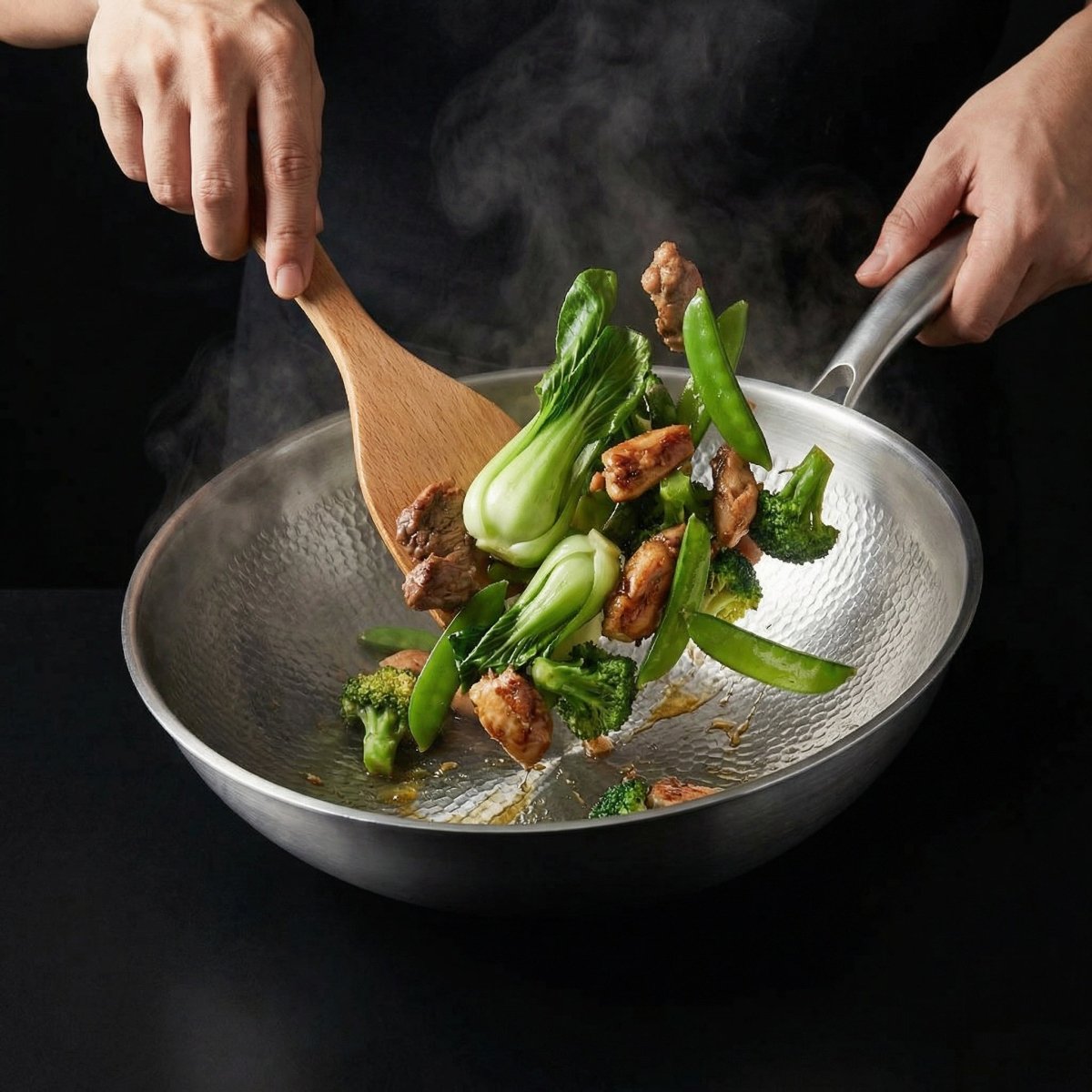 Wokpfanne aus Titan mit Pak Choi, Brokkoli, Zuckerschoten und Fleisch, von zwei Händen mit einem Holzspatel bewegt.