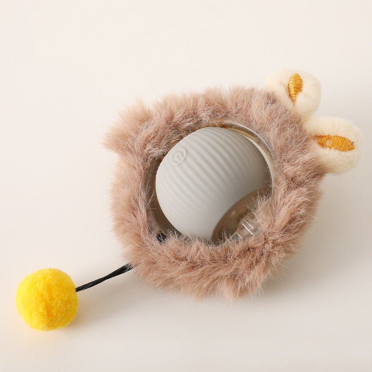 Grauer Ball in braunem Plüschmantel mit zwei cremefarbenen Öhrchen und gelbem Bommel an dünner Schnur, Draufsicht.