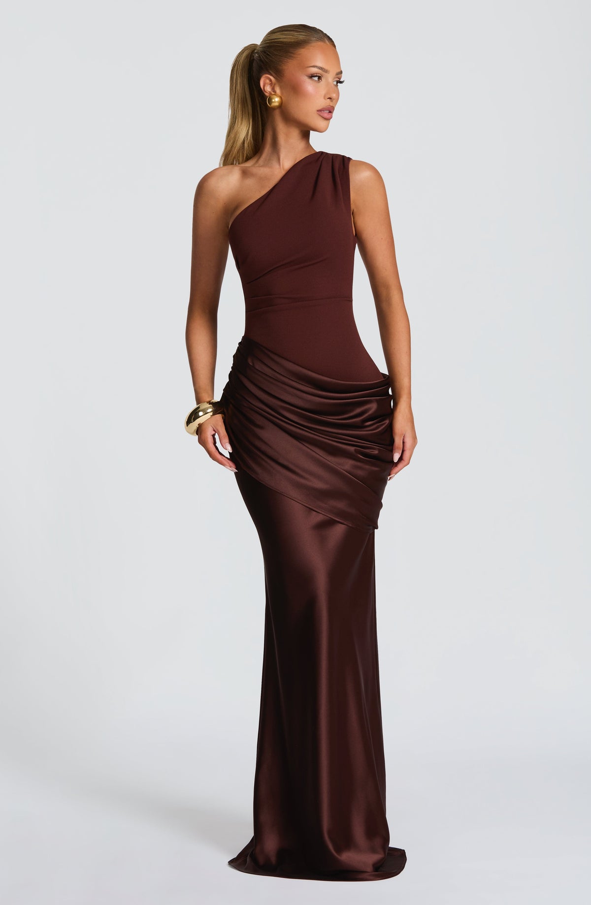 Frau mit blondem Pferdeschwanz in schokoladenbraunem One-Shoulder-Maxikleid mit glänzendem, gerafftem Satinrock, dazu goldenes Armband. 25