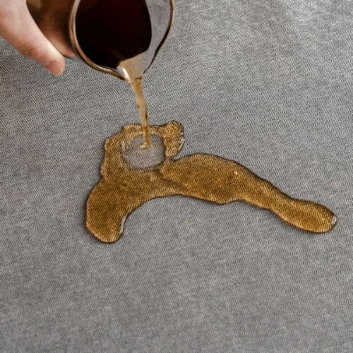 Hand gießt dunkle Flüssigkeit auf den grauen Bezug, die sich auf dem Stoff sammelt.
