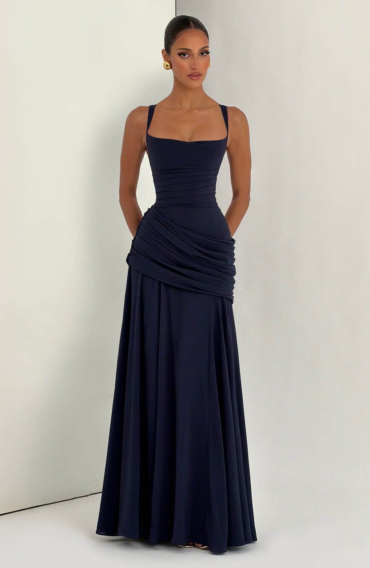 Das marineblaue Kleid vor hellem Hintergrund mit Wandecke, getragen von einem Model mit Händen in den Taschen. 11
