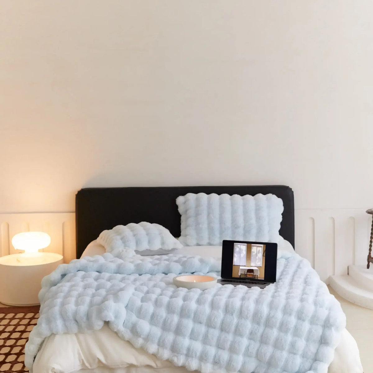 Bett mit hellblauer Noppen-Plüschdecke und passenden Kissen, darauf Laptop und Kerzenschale, Lampe links 5
