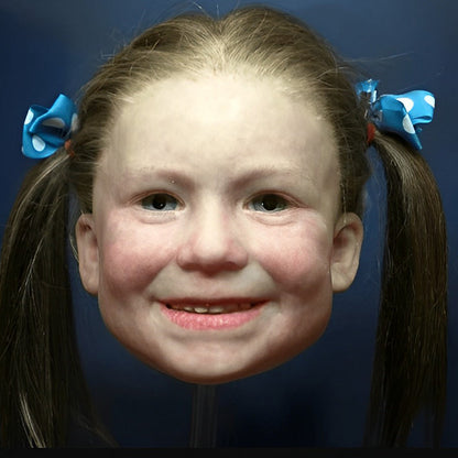 Realistische Gesichtsmaske eines lächelnden Mädchens mit braunen Zöpfen und blauen Schleifen mit weißen Punkten.
