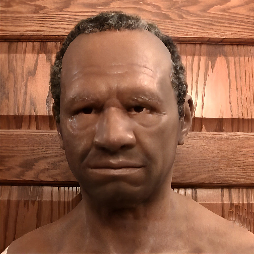 Frontalansicht einer realistischen Maske mit dunklem Hautton, kurzem gelocktem Haar und ernstem Gesichtsausdruck vor einer Holzwand. 23
