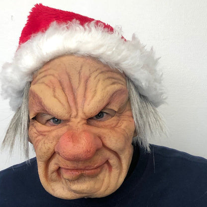 Faltige Maske eines grimmig blickenden Alten mit grauen Haarsträhnen, getragen mit roter Weihnachtsmütze mit weißem Plüschrand vor heller Wand. 24