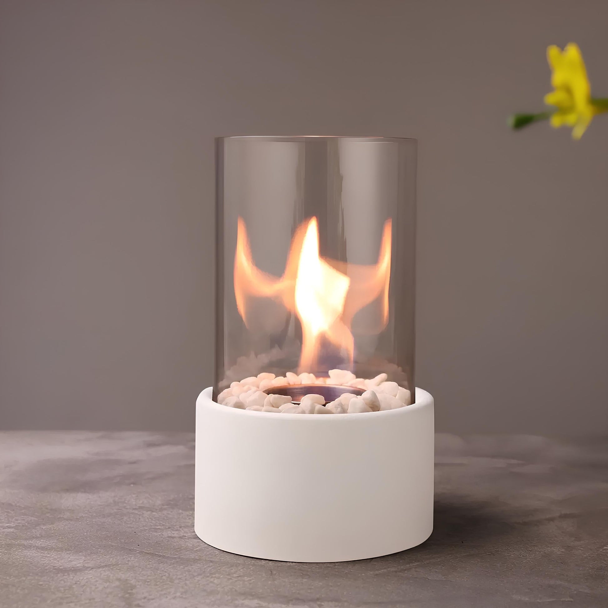 Moderner Tischkamin mit Glaszylinder, weißem Sockel und dekorativen Steinen, ideal für Innenräume.