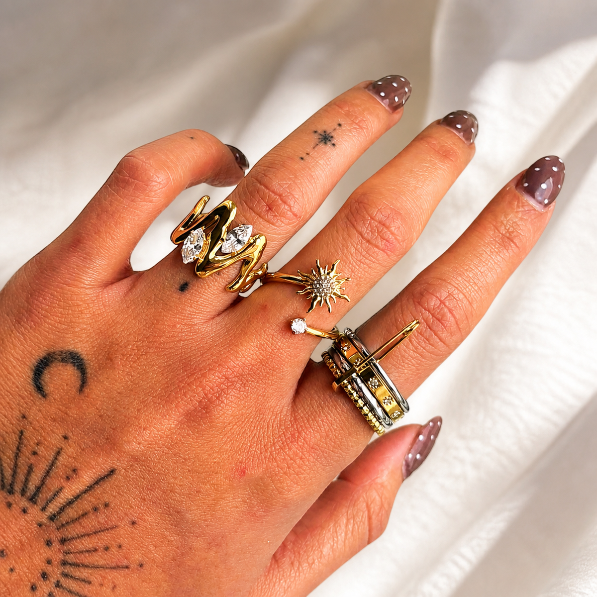 Hand mit getragenem Ring am Zeigefinger, kombiniert mit weiteren goldfarbenen Ringen, vor weißem Stoff. 3