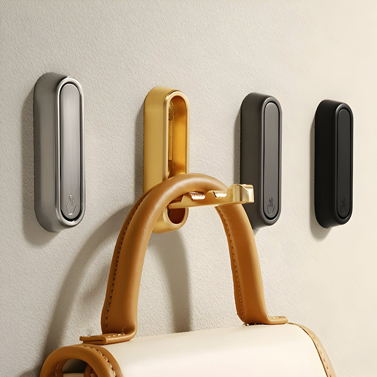 Vier ovale Klapphaken in Silber, Gold, Grau und Schwarz an einer Wand, am goldenen hängt eine braune Ledertasche.