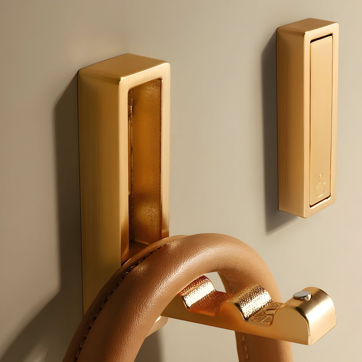 Zwei goldene Klapphaken an einer beigen Wand, einer ausgeklappt mit hellbraunem Lederhenkel, der andere flach eingeklappt.