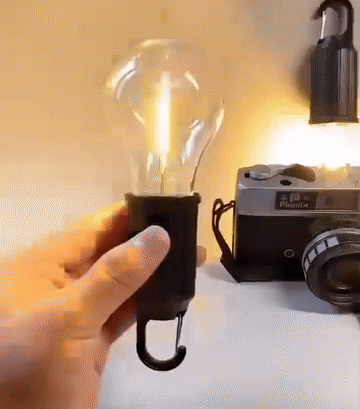 Hand hält die Lampe mit Karabiner neben einer Vintage-Kamera, im Hintergrund leuchtet eine weitere Lampe 5