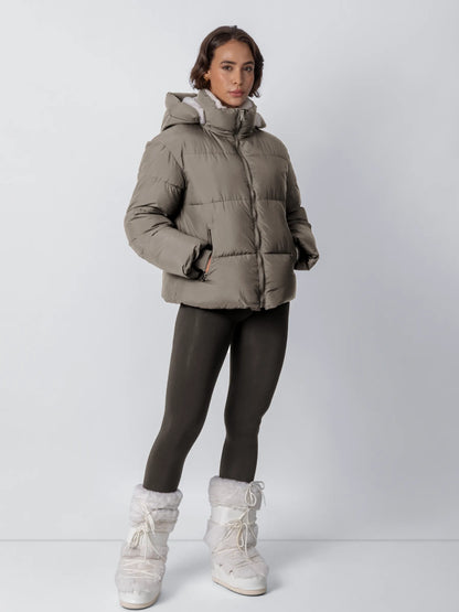 Ganzkörperansicht: Model in taupefarbener Steppjacke mit Kapuze, dunklen Leggings und weißen Plüschstiefeln, Hände in den 21