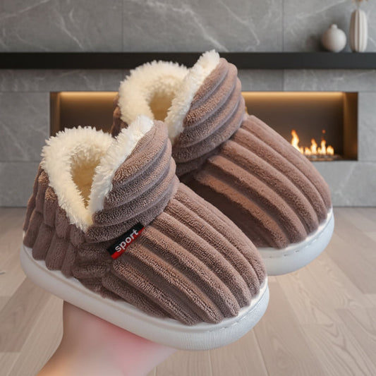 Braune gerippte Hausschuhe mit weißem Teddyfutter, von einer Hand vor einem Kamin gehalten.