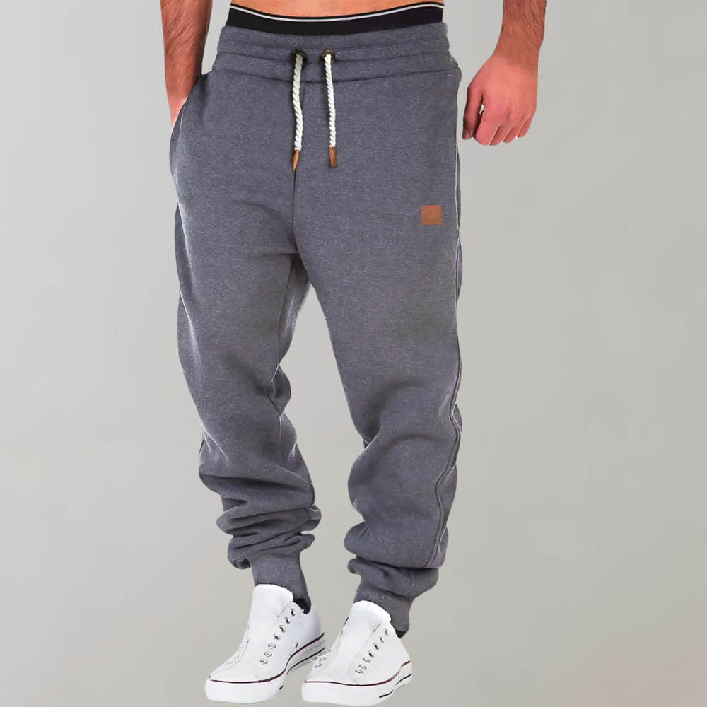 Graue Jogginghose mit hellem Kordelzug und kleinem braunem Aufnäher, von einem Model mit weißen Sneakern getragen 2