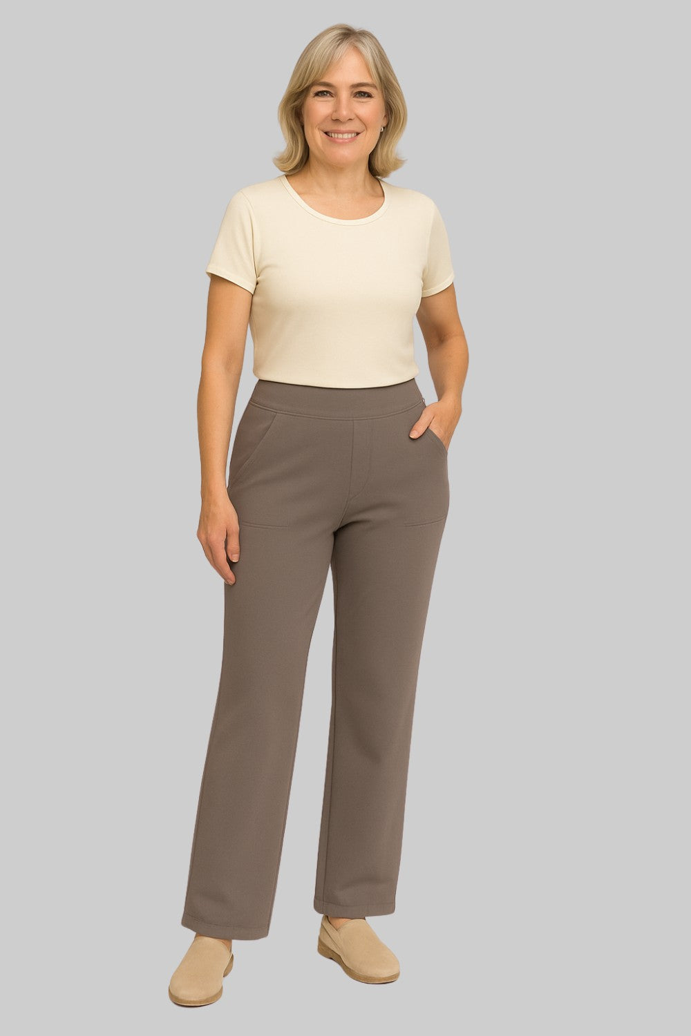 Ganzkörperbild einer lächelnden älteren Frau in taupefarbener Hose mit hohem Bund, cremefarbenem Shirt und beigen Slippern. 2
