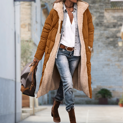 Frau trägt die lange Steppjacke in Camel offen mit weichem Teddyfutter zu Jeans, braunen Stiefeln und Handtasche vor einer Steinwand. 9