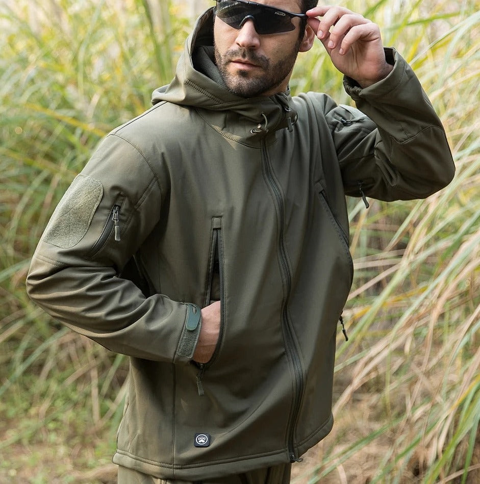 Model in olivgrüner Jacke im hohen Gras mit Sonnenbrille, eine Hand in der Tasche und sichtbarer Ärmeltasche mit Klettfläche.